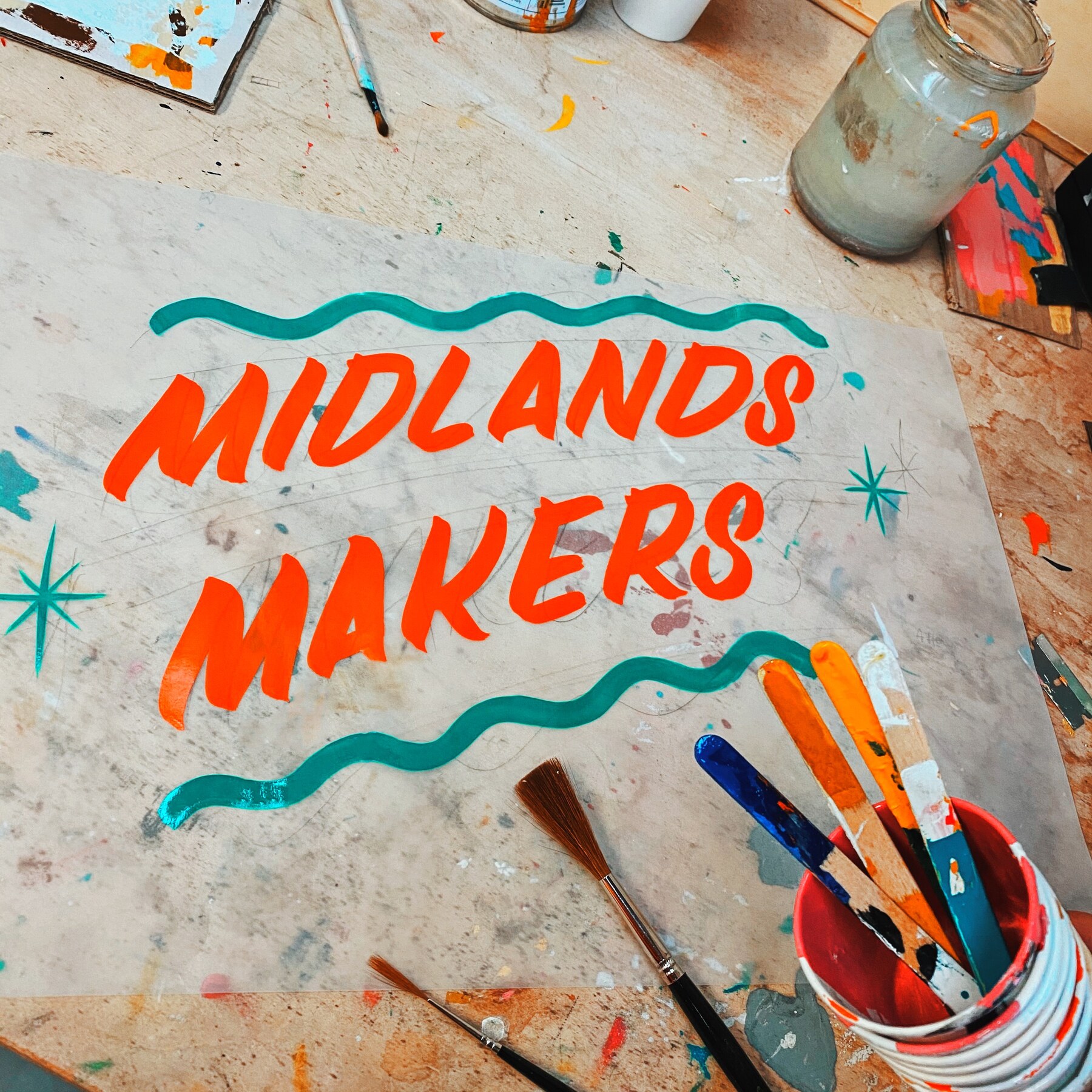 Hand-painted Midlands Makers lettering on a paint-splattered studio bench, surrounded by brushes and jars